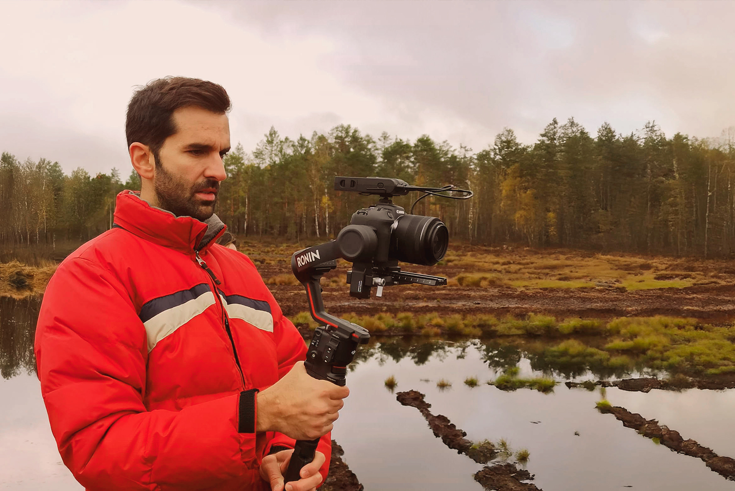 Man in a red jacket filming with a camera mounted on a gimbal beside a wetland surrounded by forest.